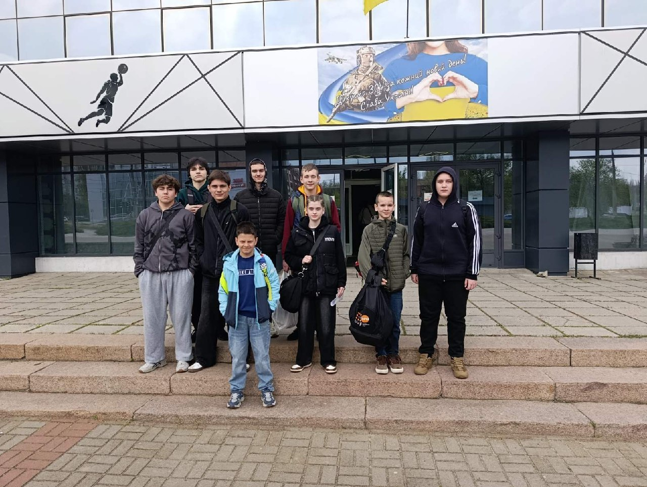 Група підлітків у спортивному та повсякденному одязі стоїть перед входом до сучасної будівлі. Над дверима розміщено патріотичний плакат, а на фасаді видно силует баскетболіста.