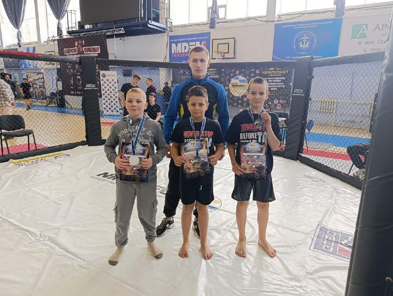 Троє юних спортсменів із дипломами та медалями стоять усередині клітки для єдиноборств. Позаду них стоїть тренер або старший наставник. На задньому плані видно облаштування спортивного залу з банерами змагань та баскетбольним кільцем.