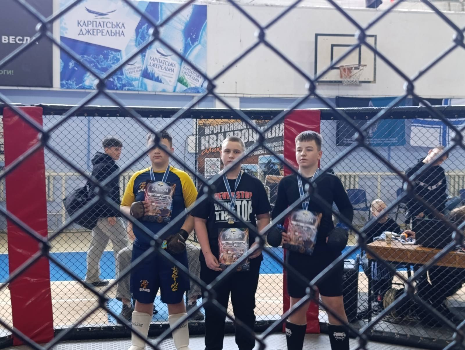 Троє юних спортсменів із медалями та дипломами в руках стоять усередині клітки для поєдинків. Фото зняте крізь металеву сітку огорожі. На задньому плані спортивного залу видно баскетбольне кільце та рекламні банери.
