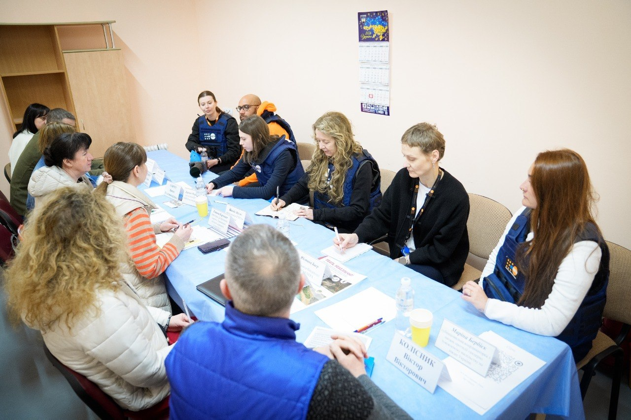 Робоча зустріч за довгим столом у приміщенні. Частина присутніх у синіх жилетах гуманітарних організацій спілкується з іншими учасниками. На столі лежать блокноти, ручки та таблички з іменами.