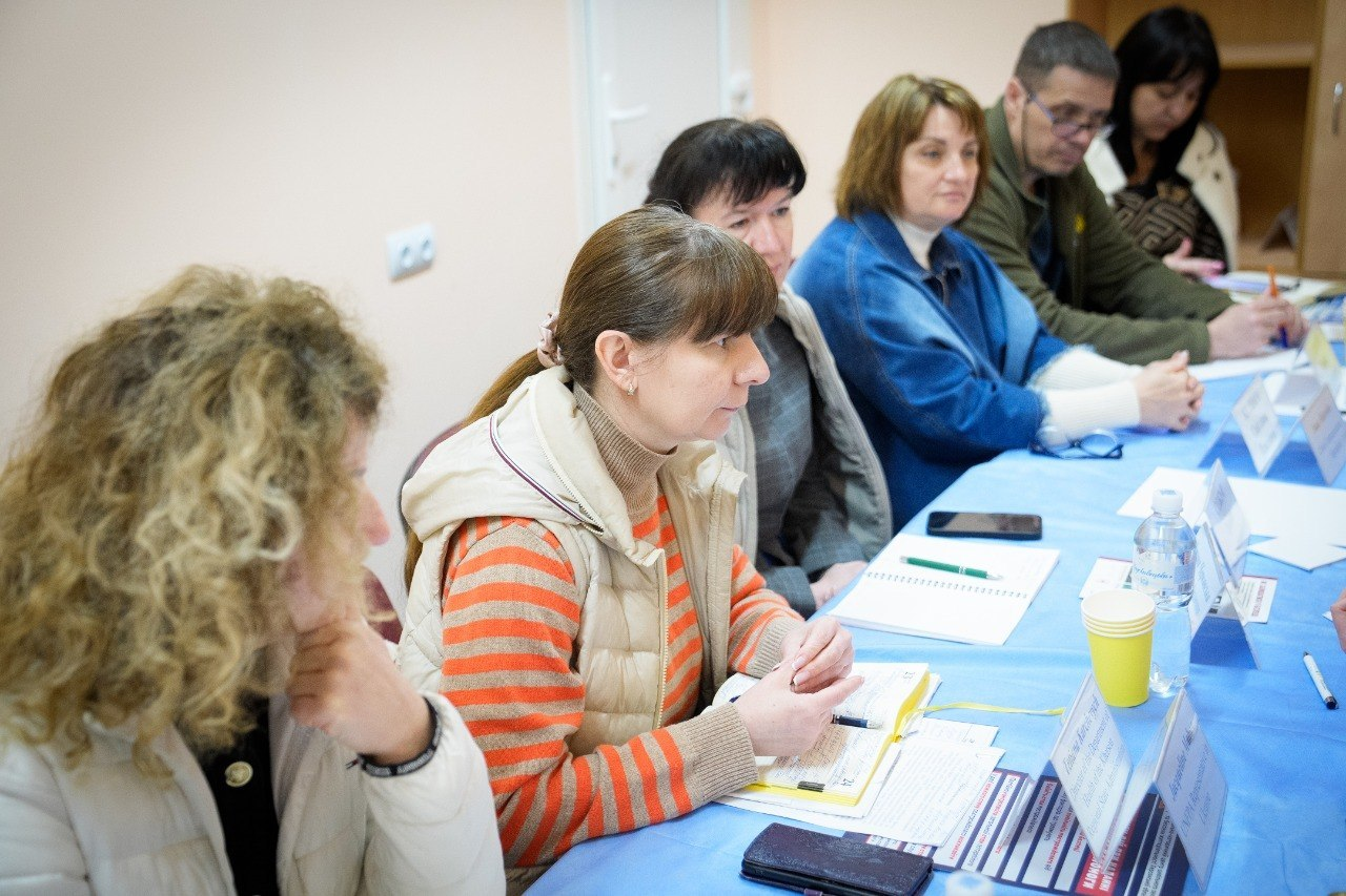 Група людей сидить у ряд за столом із блакитною скатертиною під час наради чи тренінгу. Учасники зосереджено слухають, перед ними лежать відкриті блокноти для записів, ручки, смартфони та інформаційні таблички.