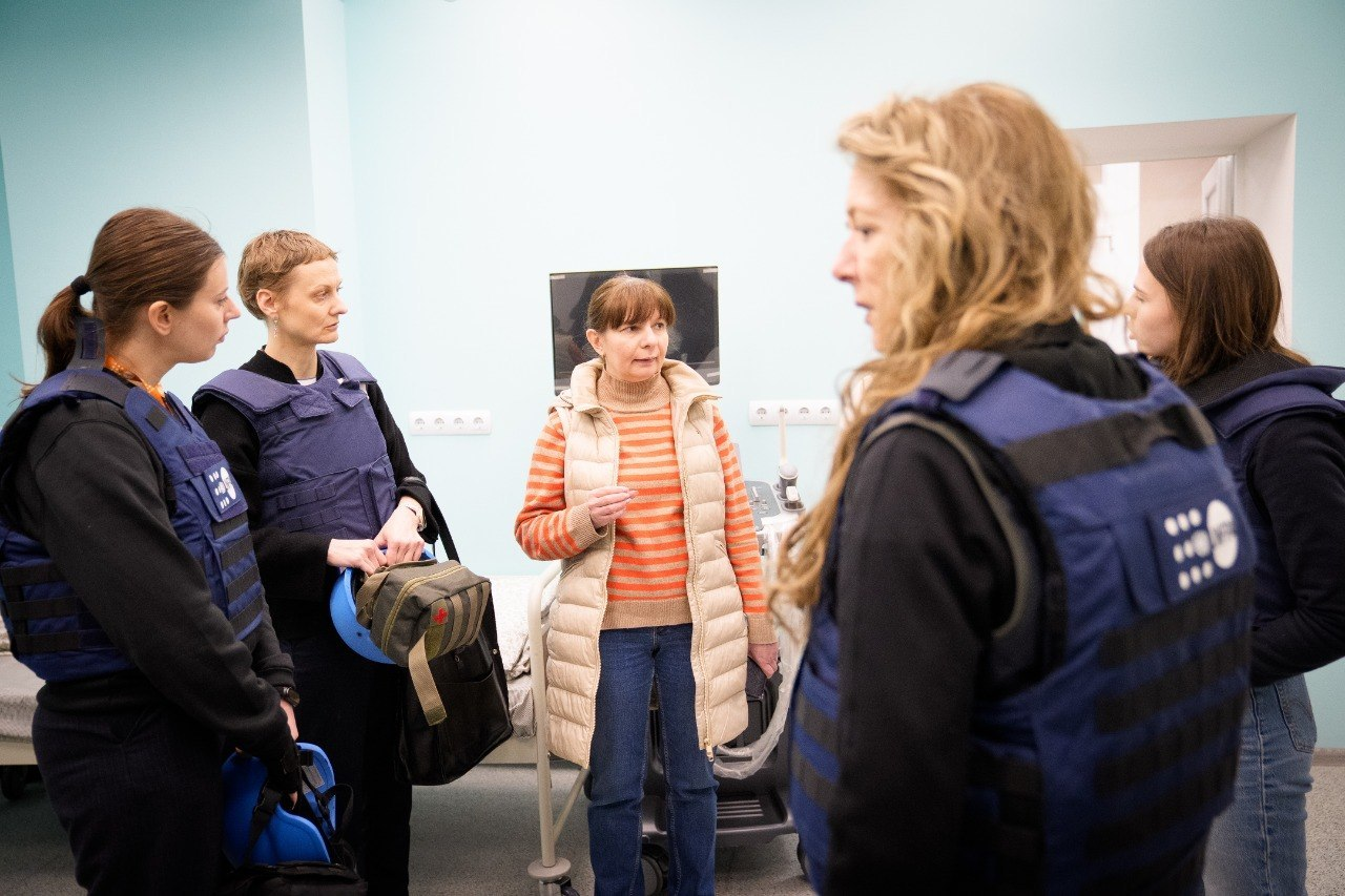 Група людей у світлому приміщенні медичного або соціального призначення. У центрі жінка розповідає щось відвідувачам, частина з яких одягнена в сині захисні жилети та тримає шоломи. На задньому плані видно медичне обладнання та кушетку.