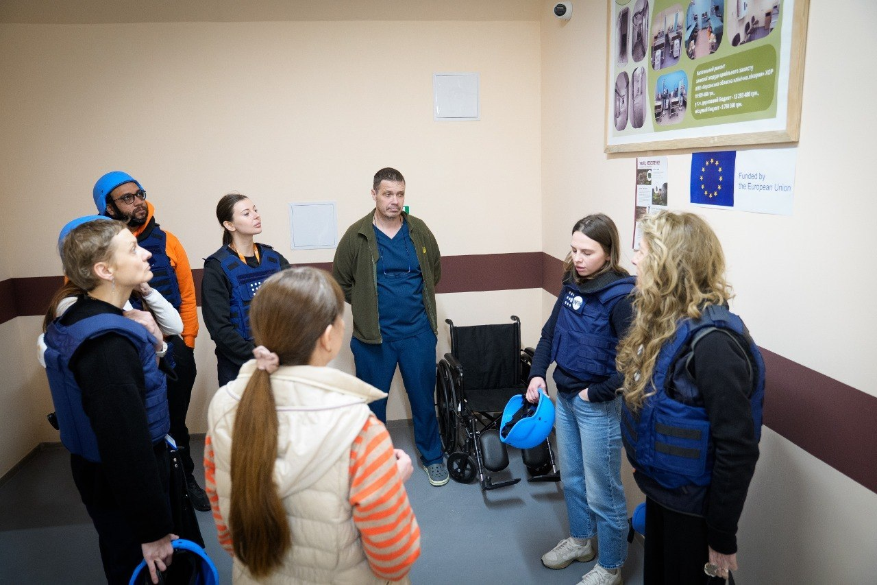 Група людей у приміщенні біля інформаційного стенда з прапором ЄС та інвалідного візка. Більшість присутніх одягнені в сині захисні жилети та тримають шоломи, уважно слухаючи чоловіка в медичній формі.