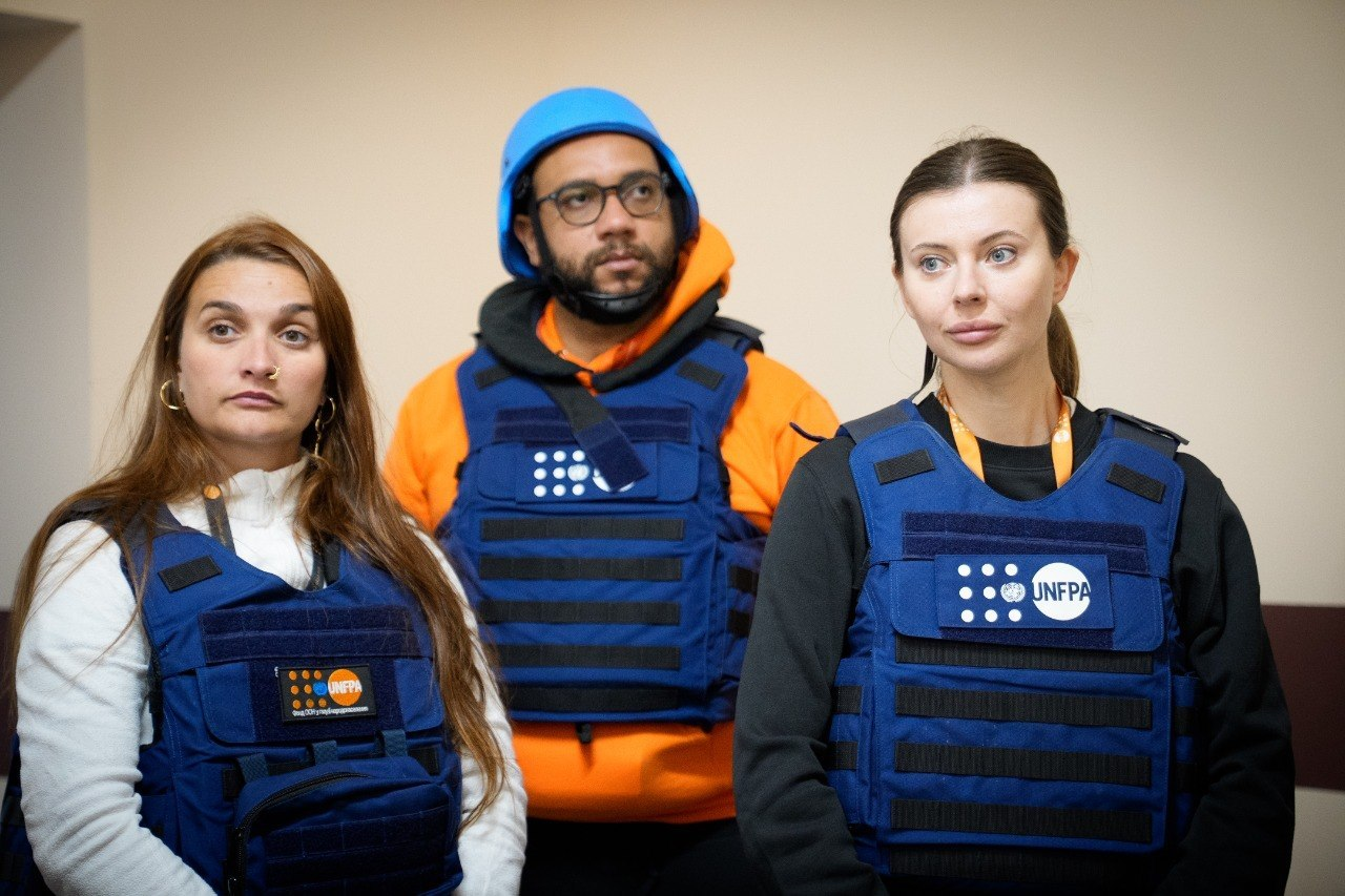Троє представників міжнародної організації UNFPA у синіх захисних жилетах. Чоловік у центрі також має блакитний шолом. Вони стоять поруч і серйозно дивляться перед собою, наче під час офіційного візиту чи прослуховування звіту.