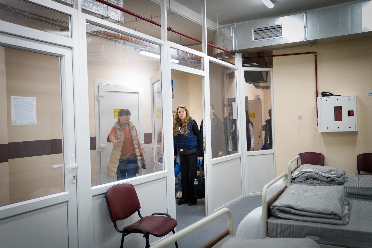 Група людей оглядає приміщення з ліжками та скляними перегородками. У коридорі за дверима видно представницю гуманітарної місії в синьому жилеті під час обговорення.