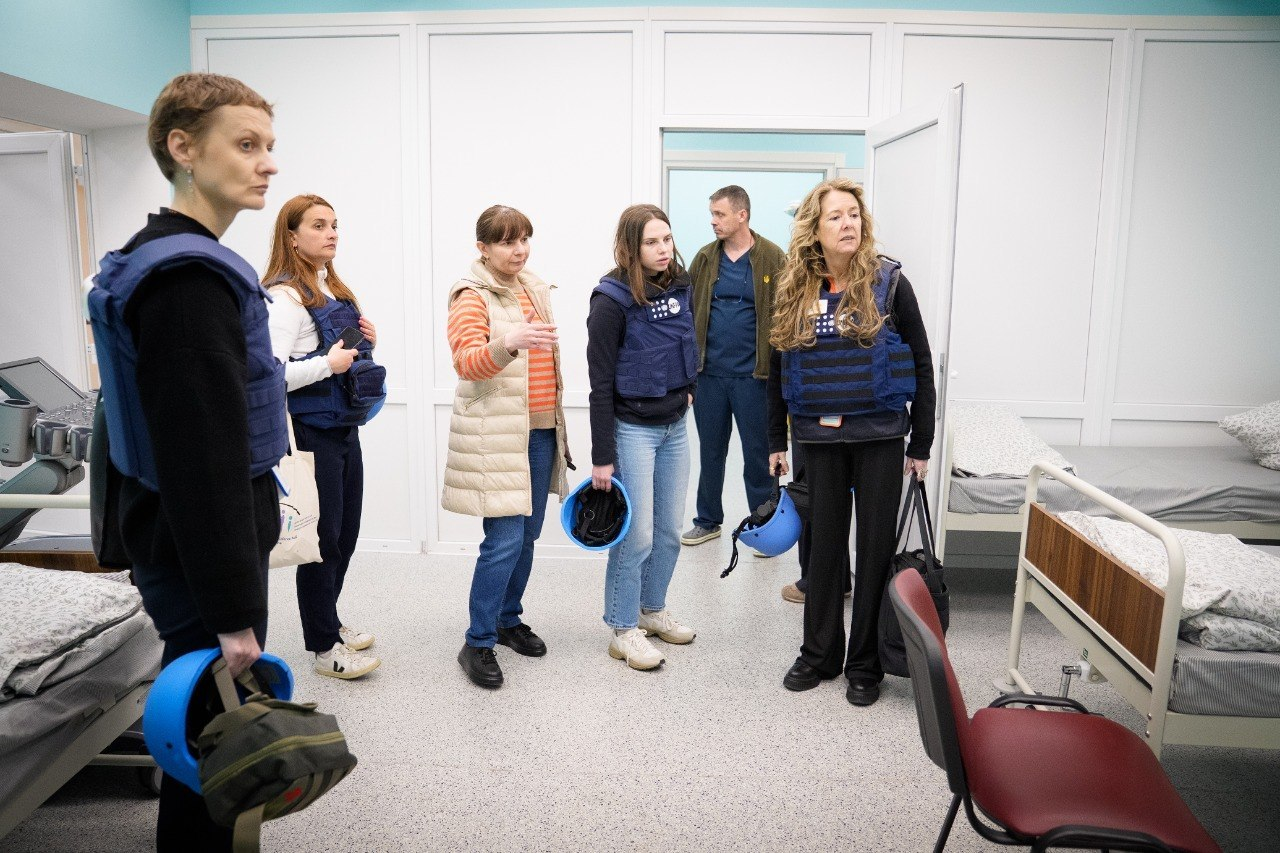 Група людей у світлій медичній палаті з ліжками. Кілька жінок одягнені в сині захисні жилети та тримають шоломи, уважно слухаючи представницю закладу. На задньому плані в дверях стоїть чоловік у медичній формі.