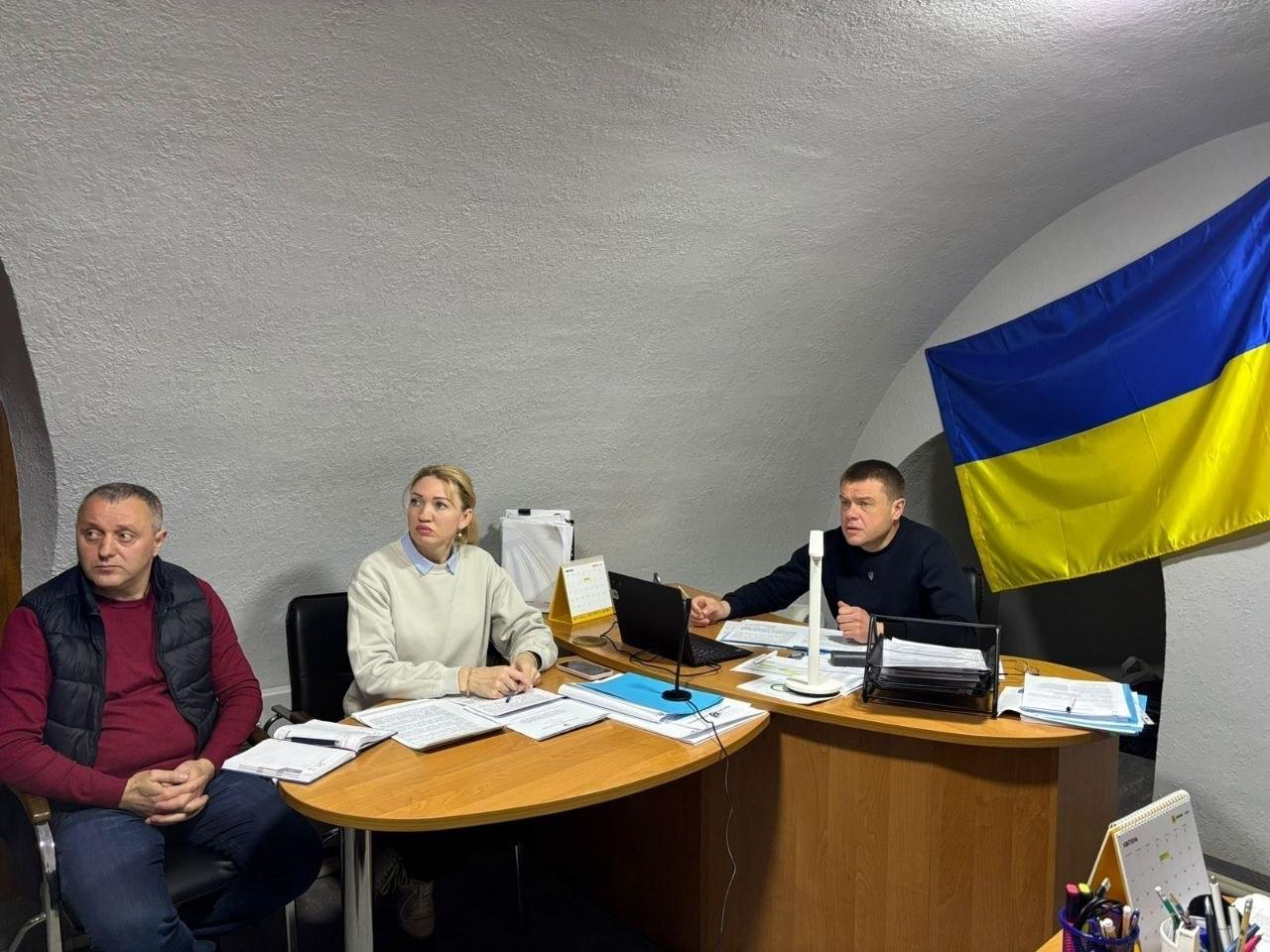 Троє людей сидять за робочими столами в приміщенні зі склепінчастою стелею. На столах лежать документи та ноутбук, а на стіні праворуч закріплений прапор України. Учасники зустрічі зосереджено дивляться в одному напрямку, наче під час наради або відеодзвінка.