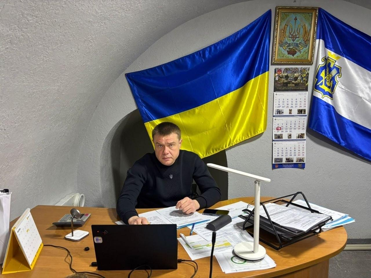 Чоловік у чорному светрі сидить за робочим столом у приміщенні зі склепінчастою стелею. Перед ним відкритий ноутбук, лампа та стоси документів. На стіні позаду розміщені прапор України, ще один синьо-білий прапор та календарі.