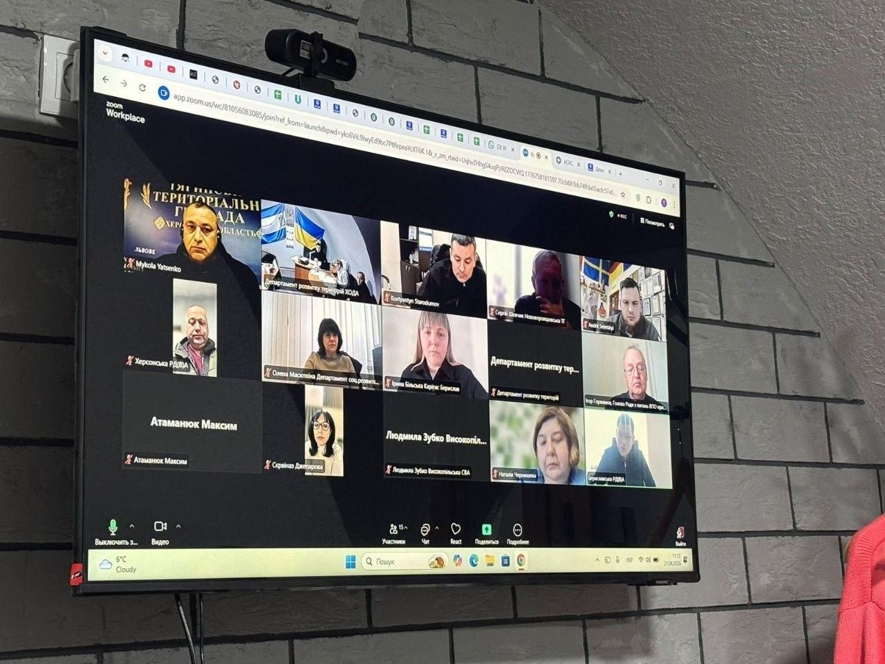 Великий телевізійний екран, закріплений на сірій цегляній стіні, на якому транслюється онлайн-нарада в Zoom. У сітці відеозв'язку видно багато учасників, деякі вікна підписані як офіційні департаменти та громади Херсонщини. Зверху на моніторі встановлена веб-камера.