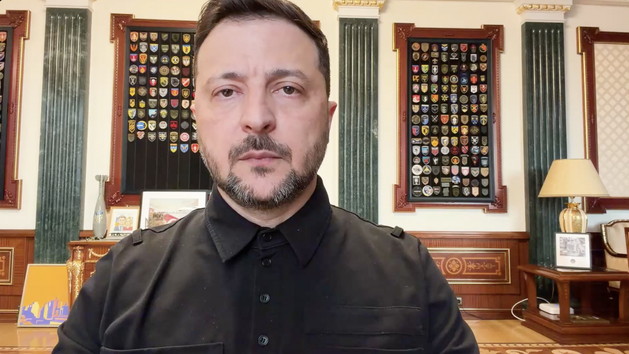 Чоловік у чорній сорочці на фоні офіційного кабінету. На стіні за ним розміщені дві великі дошки з численними військовими шевронами та нашивками.