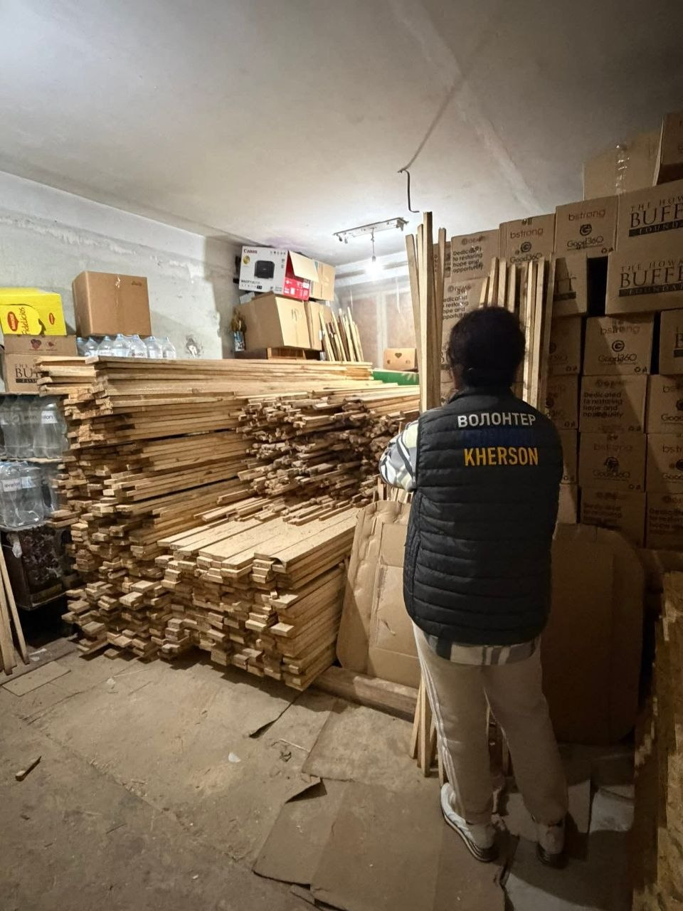 Волонтер у жилеті з написом «Волонтер Kherson» працює на складі гуманітарної допомоги. У приміщенні складовано велику кількість дерев'яних рейок, бутлі з водою та картонні коробки з вантажем.