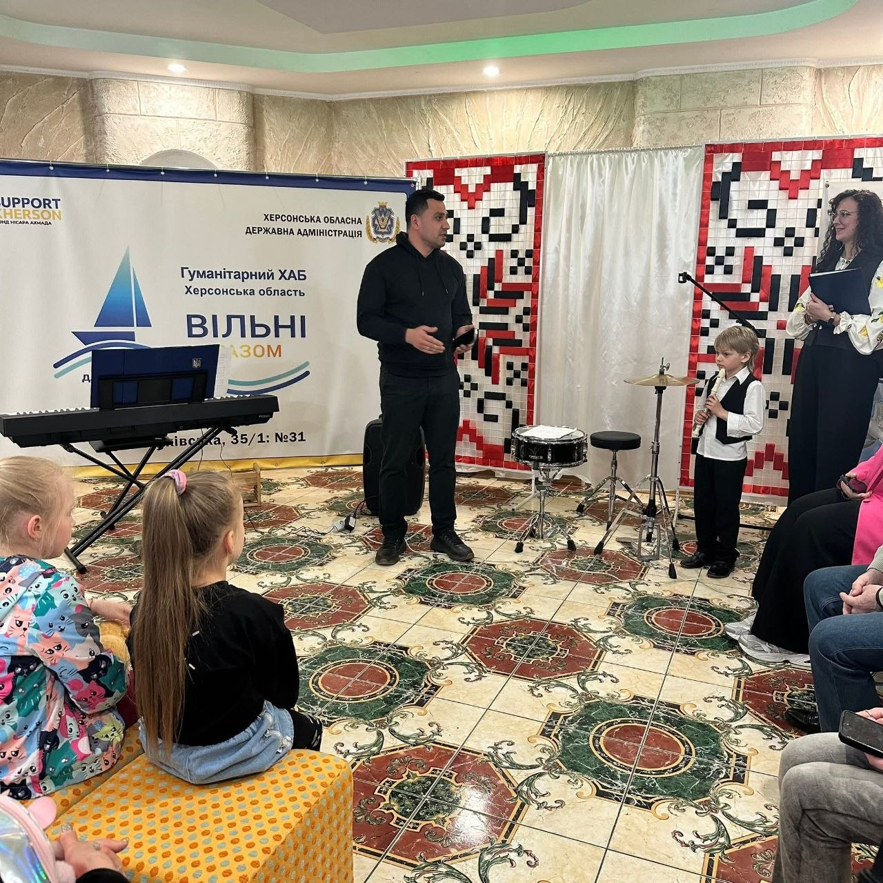 Захід у гуманітарному хабі Херсонської області: чоловік виступає перед аудиторією в залі, поруч стоїть дитина з музичним інструментом. У приміщенні встановлено банери організацій, музичне обладнання та декоративні панно з українським орнаментом.