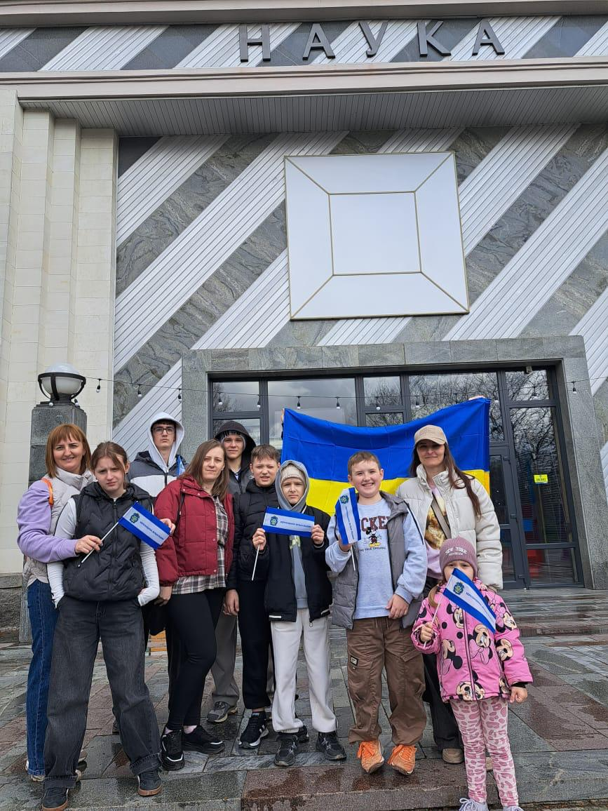 Група дітей та дорослих стоїть перед входом до будівлі з написом «НАУКА». Учасники тримають великий прапор України та маленькі синьо-білі прапорці, позуючи для спільного фото на фоні сучасного фасаду.