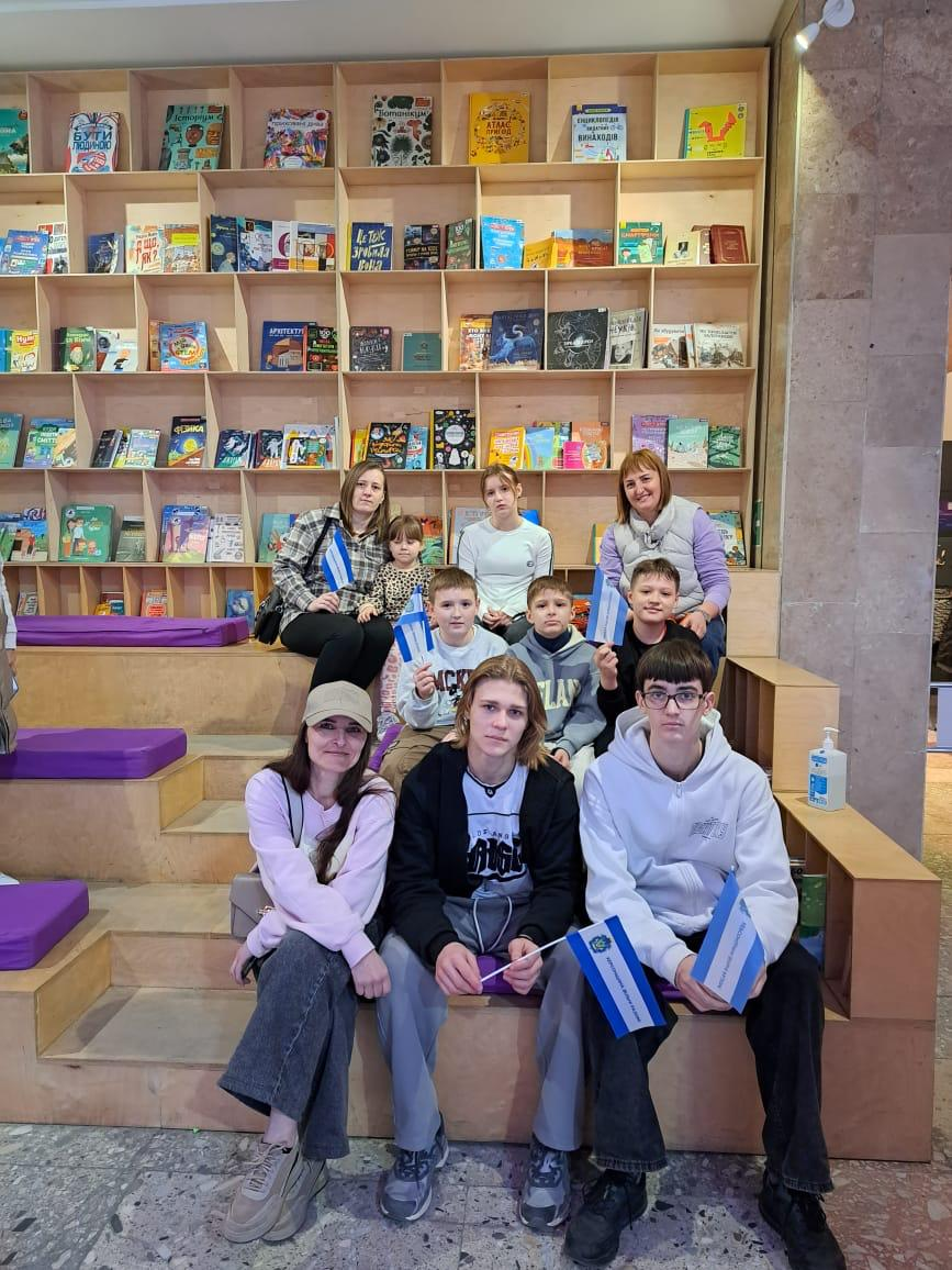 Група дітей, підлітків та дорослих позирує для спільного фото в сучасному просторі з великим стелажем дитячих книжок. Деякі учасники тримають у руках невеликі синьо-білі прапорці, сидячи на дерев’яних сходах-подіумах із фіолетовими подушками.