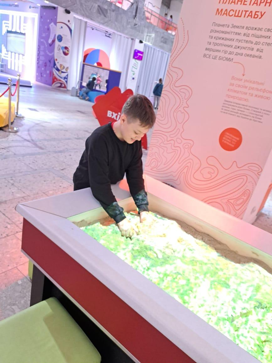 Хлопець взаємодіє з інтерактивною пісочницею на науковій виставці, де світлові проекції на піску імітують топографію. На фоні видно великий інформаційний стенд «Планетарний масштаб» про різноманіття біомів Землі.