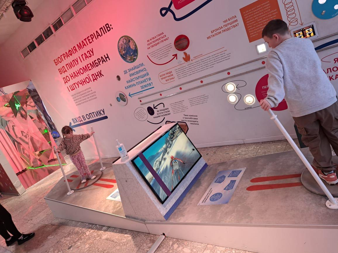Діти взаємодіють з інтерактивними експонатами на науковій виставці «Біографія матеріалів». У залі представлені інформаційні стенди з текстами та графіками, а також ігрова зона з екраном, де хлопчик та дівчинка випробують симулятор, тримаючись за поручні.