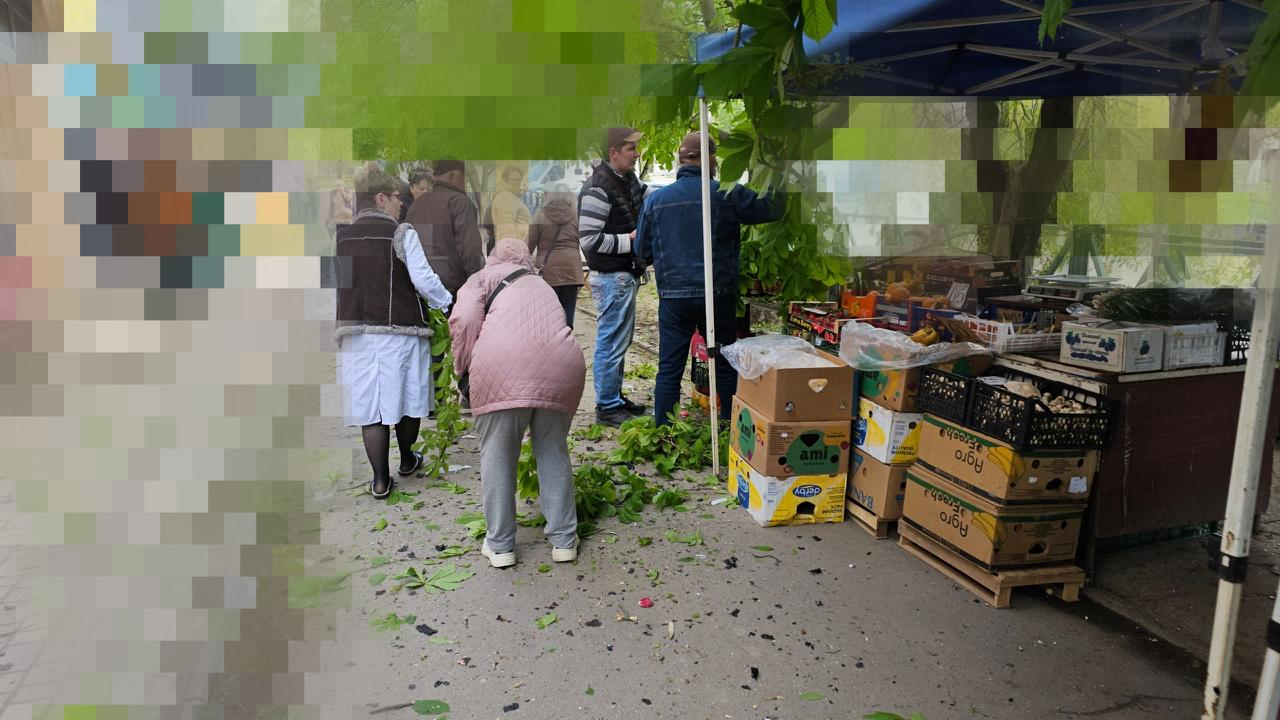 Це вулична торговельна точка під синім тентом з овочами та фруктами у ящиках. Частина зображення зліва та по центру сильно розмита (пікселізована), що приховує деталі та обличчя людей, які стоять поруч із прилавком.