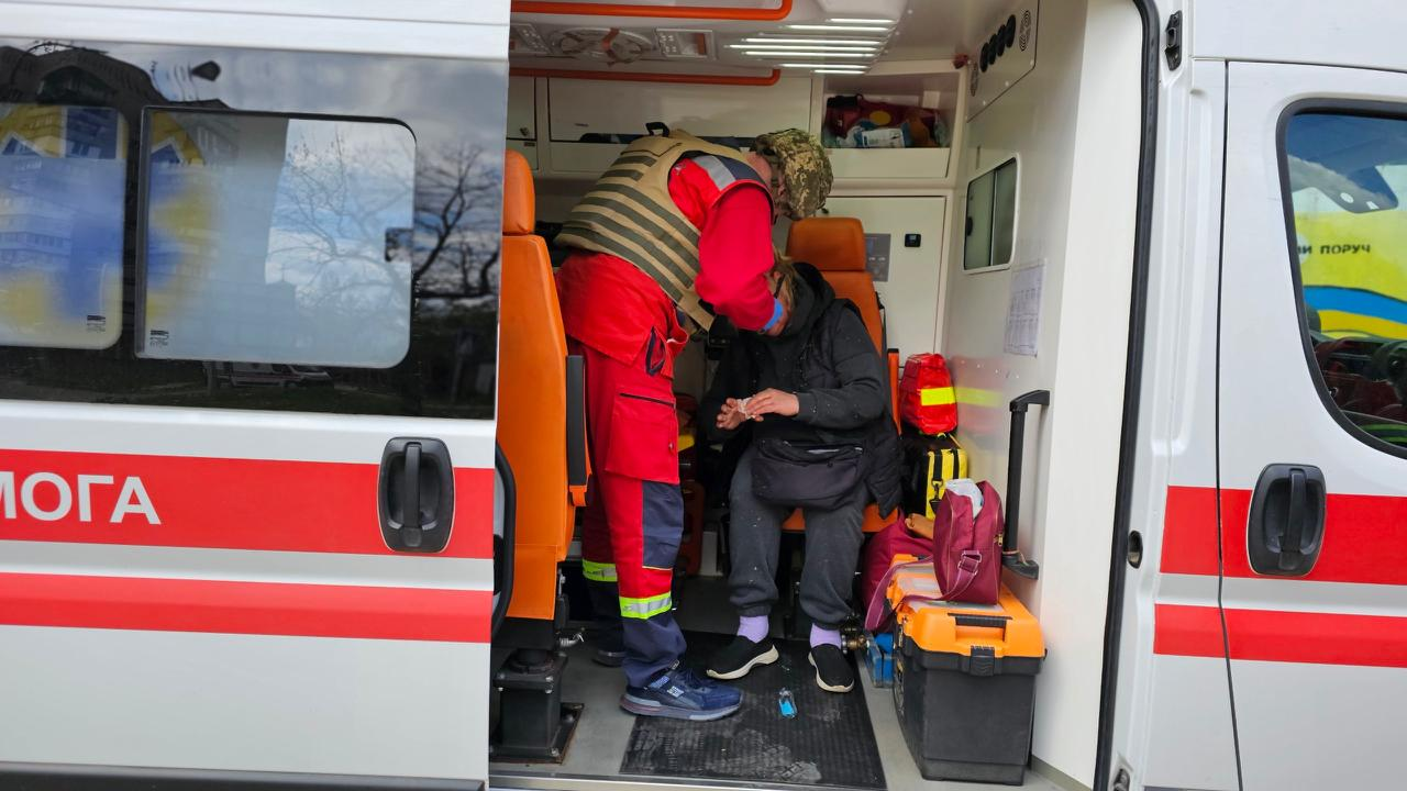 Медик у червоній уніформі та бронежилеті надає допомогу пацієнту всередині сучасного автомобіля швидкої допомоги. Салон обладнаний помаранчевими сидіннями, медичними сумками та інвентарем, а на розсувних дверях видно частину напису «ДОПОМОГА».