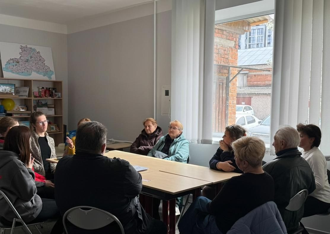 Робоча зустріч у світлому кабінеті: група людей різного віку сидить навколо столу та веде обговорення. На стіні позаду видно мапу Херсонської області, а поруч — стелаж із книжками та документами.