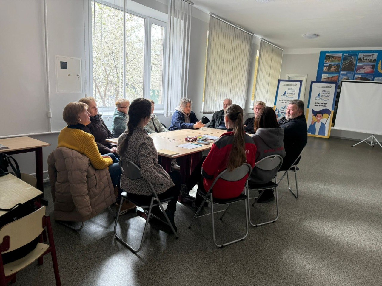 Група людей сидить за великим столом у світлому навчальному або офісному приміщенні під час робочої зустрічі. У залі встановлені інформаційні стенди з логотипами організацій, а на фоні видно велике вікно та екран для презентацій.