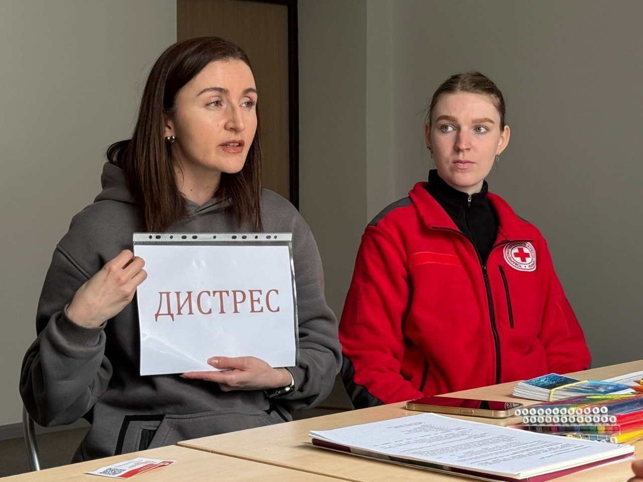 Дві жінки проводять заняття або презентацію: одна з них тримає аркуш із написом «ДИСТРЕС», інша — у червоній кофті з емблемою Червоного Хреста — сидить поруч. На столі перед ними лежать навчальні матеріали та олівці.