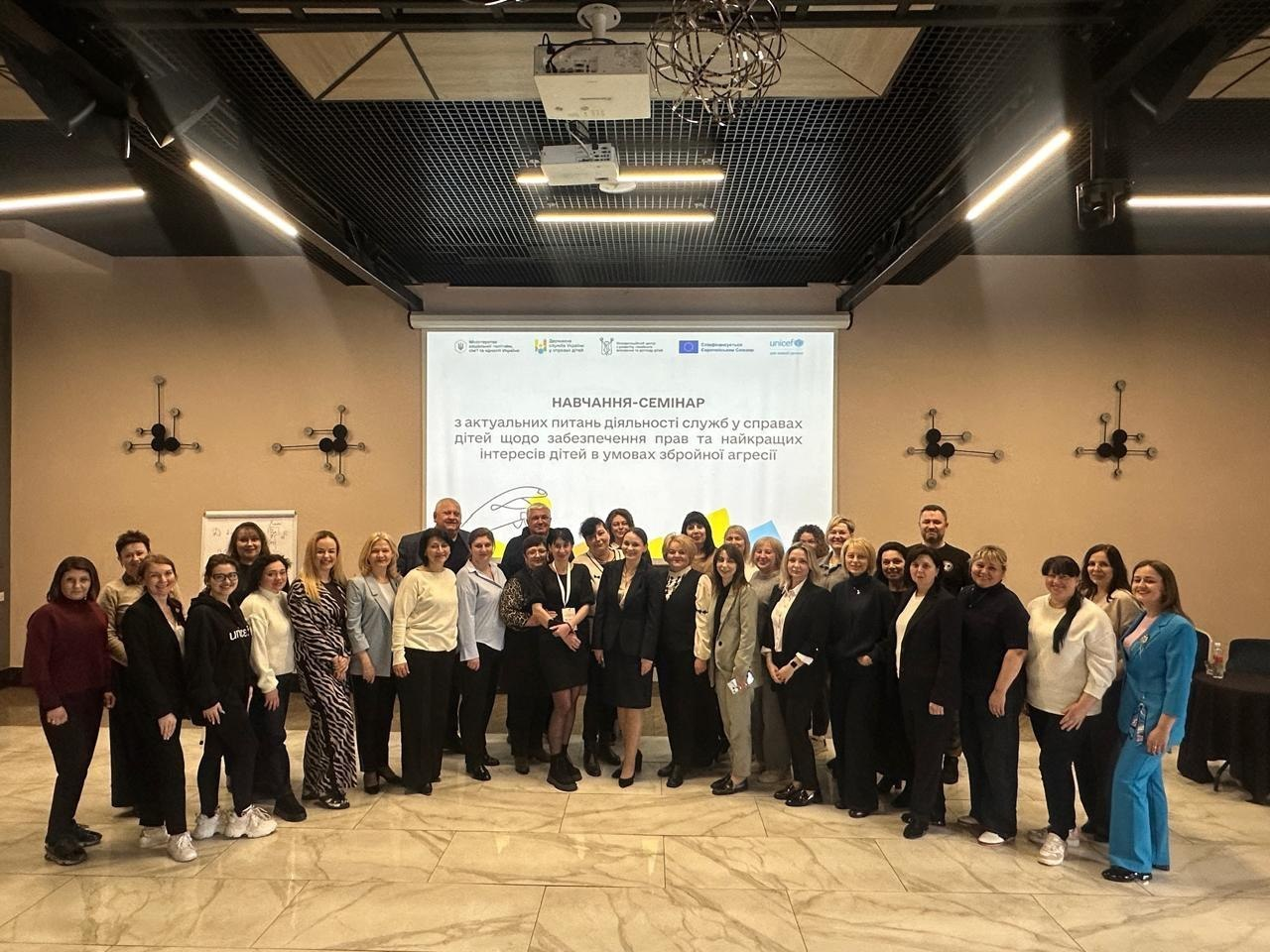 Групова фотографія учасників та учасниць навчання-семінару в конференц-залі. Велика група людей стоїть перед мультимедійним екраном із заголовком про діяльність служб у справах дітей та забезпечення прав дитини в умовах збройної агресії.