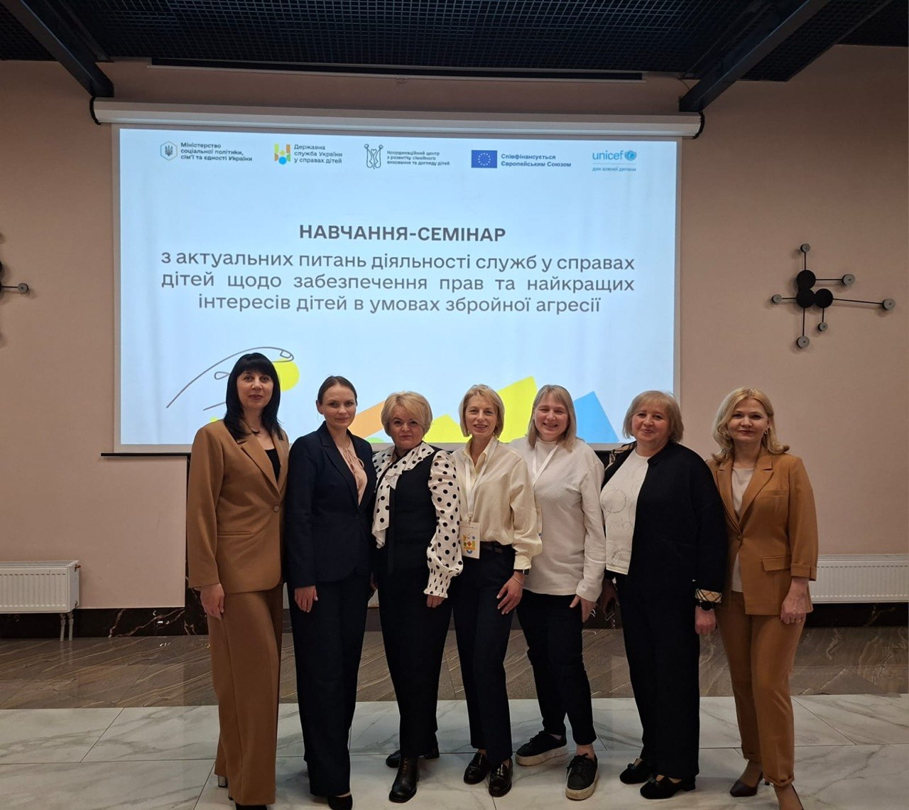 Група з семи жінок позує для спільного фото в залі на фоні мультимедійного екрана. На екрані відображено заголовок: «Навчання-семінар з актуальних питань діяльності служб у справах дітей» та логотипи організацій-партнерів, серед яких UNICEF та Мінсоцполітики.