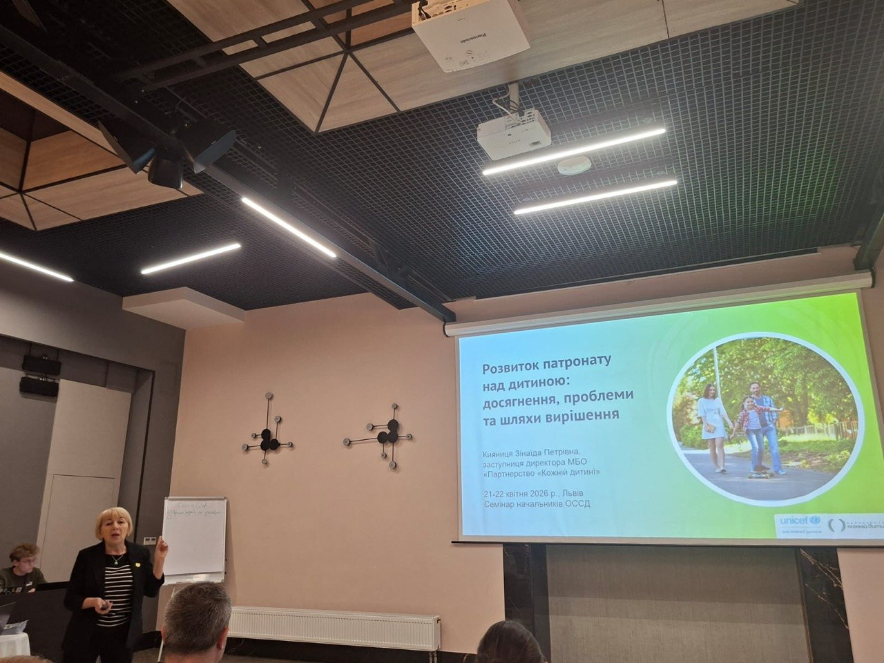 Виступ спікерки на семінарі в конференц-залі. На мультимедійному екрані відображено слайд на тему розвитку патронату над дитиною з вказаною датою проведення заходу у квітні 2026 року у Львові.