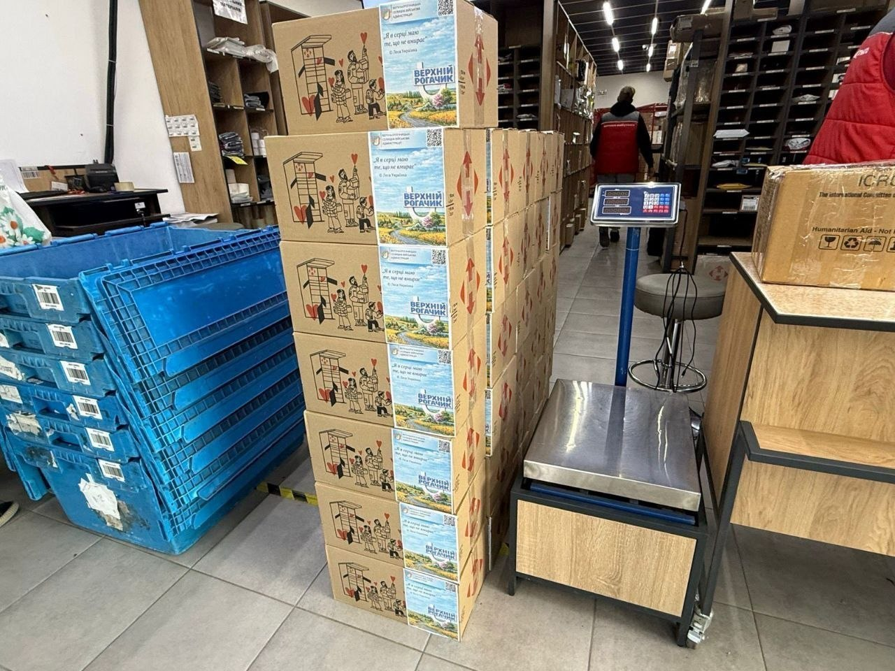 Складське приміщення з гуманітарною допомогою: у центрі стоїть висока стопка картонних коробок із маркуванням «Верхній Рогачик». Поруч розташовані великі сині пластикові контейнери, підлогові електронні ваги та стелажі з товарами на фоні.