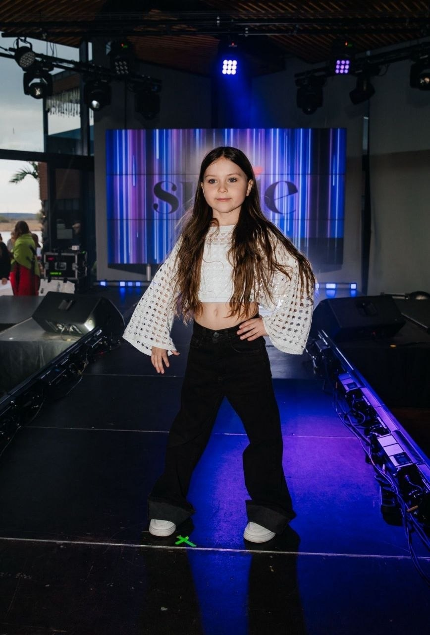 Дівчинка-модель у білому мереживному топі та чорних штанах позує на подіумі під час показу мод.