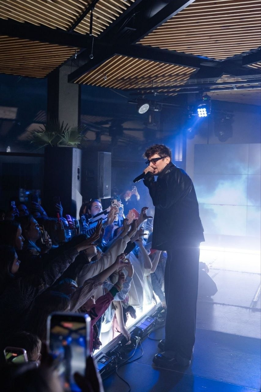 Виконавець у темних окулярах співає на сцені перед натовпом фанатів у світлі софітів.