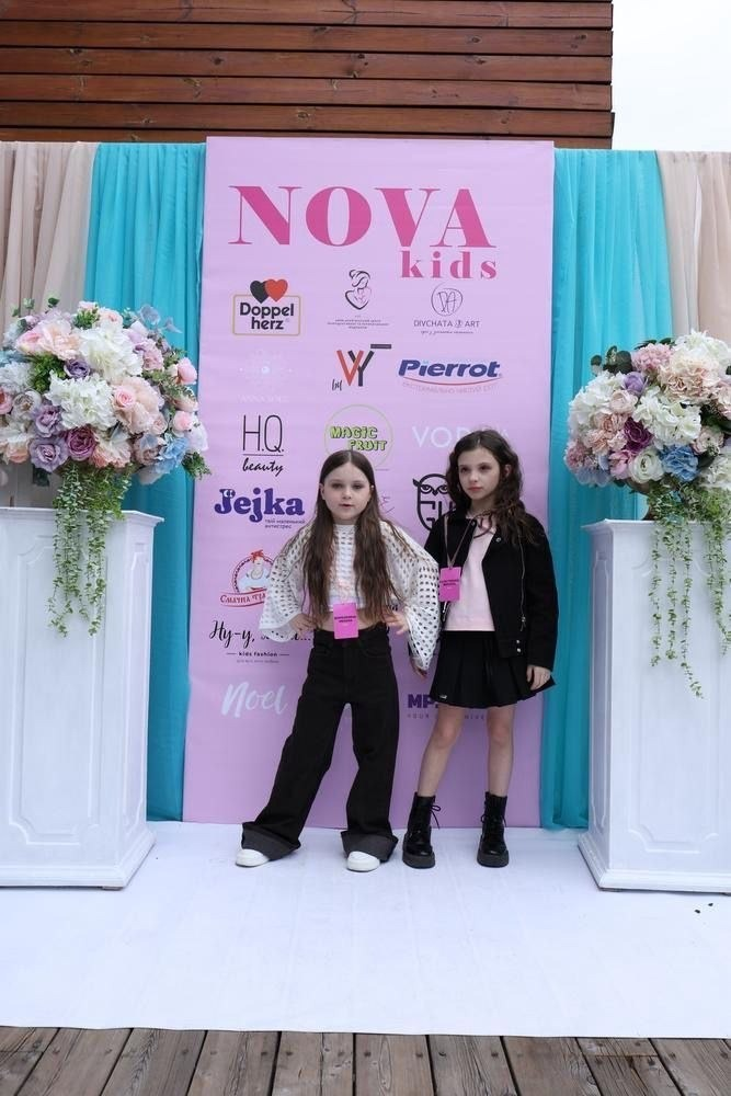 Дві дівчинки-моделі позують на фоні рожевого банера з логотипами біля квіткових композицій.