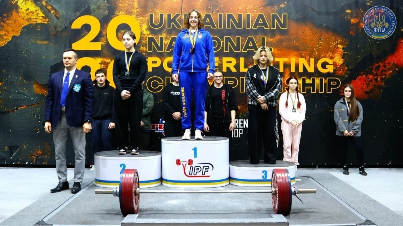 Церемонія нагородження на чемпіонаті України з пауерліфтингу. Переможці з медалями стоять на п'єдесталі на фоні офіційного банера змагань. Попереду на платформі лежить штанга.