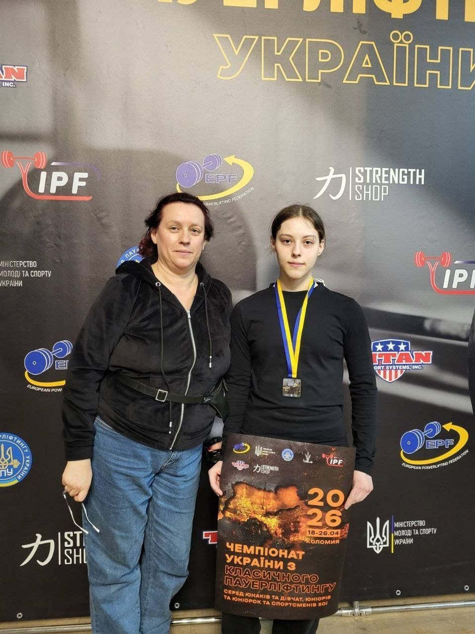 Молода спортсменка з медаллю на шиї та жінка стоять поруч на фоні банера чемпіонату України з класичного пауерліфтингу 2026. Дівчина тримає в руках афішу змагань із логотипами спортивних федерацій та партнерів.