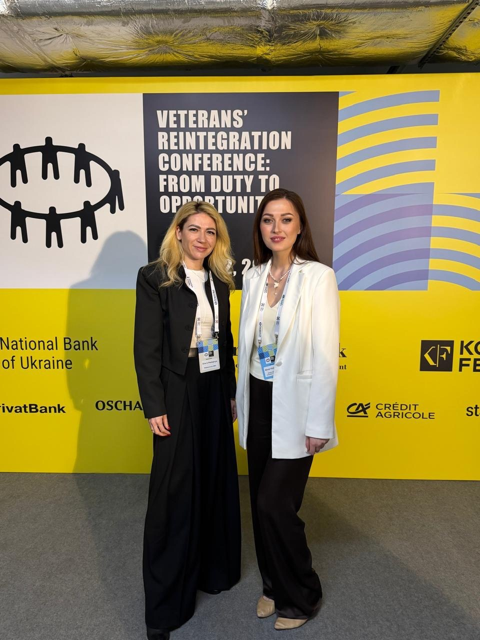 Дві жінки на фоні банера конференції з реінтеграції ветеранів. На банері видно назву заходу «Veterans’ Reintegration Conference» та логотипи партнерів, серед яких Національний банк України та інші банки.
