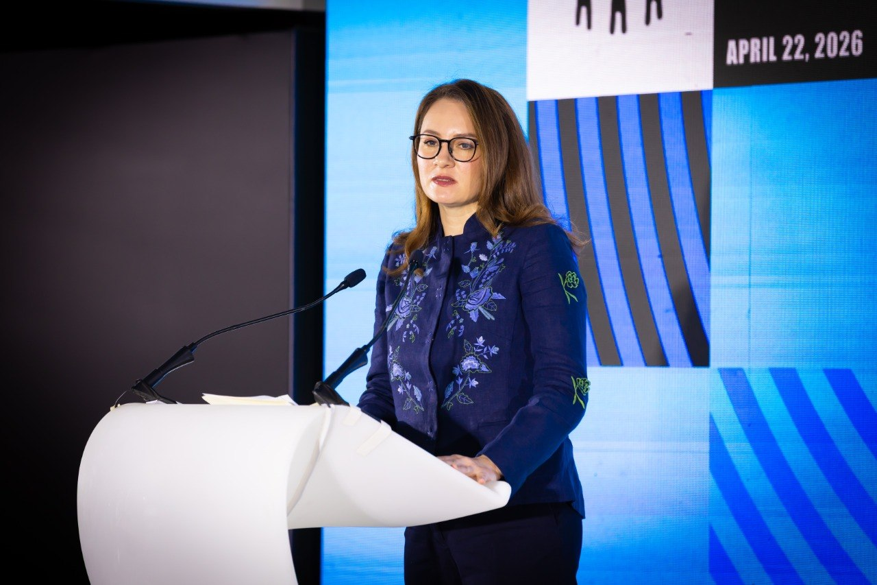 Жінка в окулярах та темно-синьому жакеті з вишивкою виступає за трибуною з мікрофонами. На задньому плані — екран із графічними елементами та датою «22 квітня 2026 року».