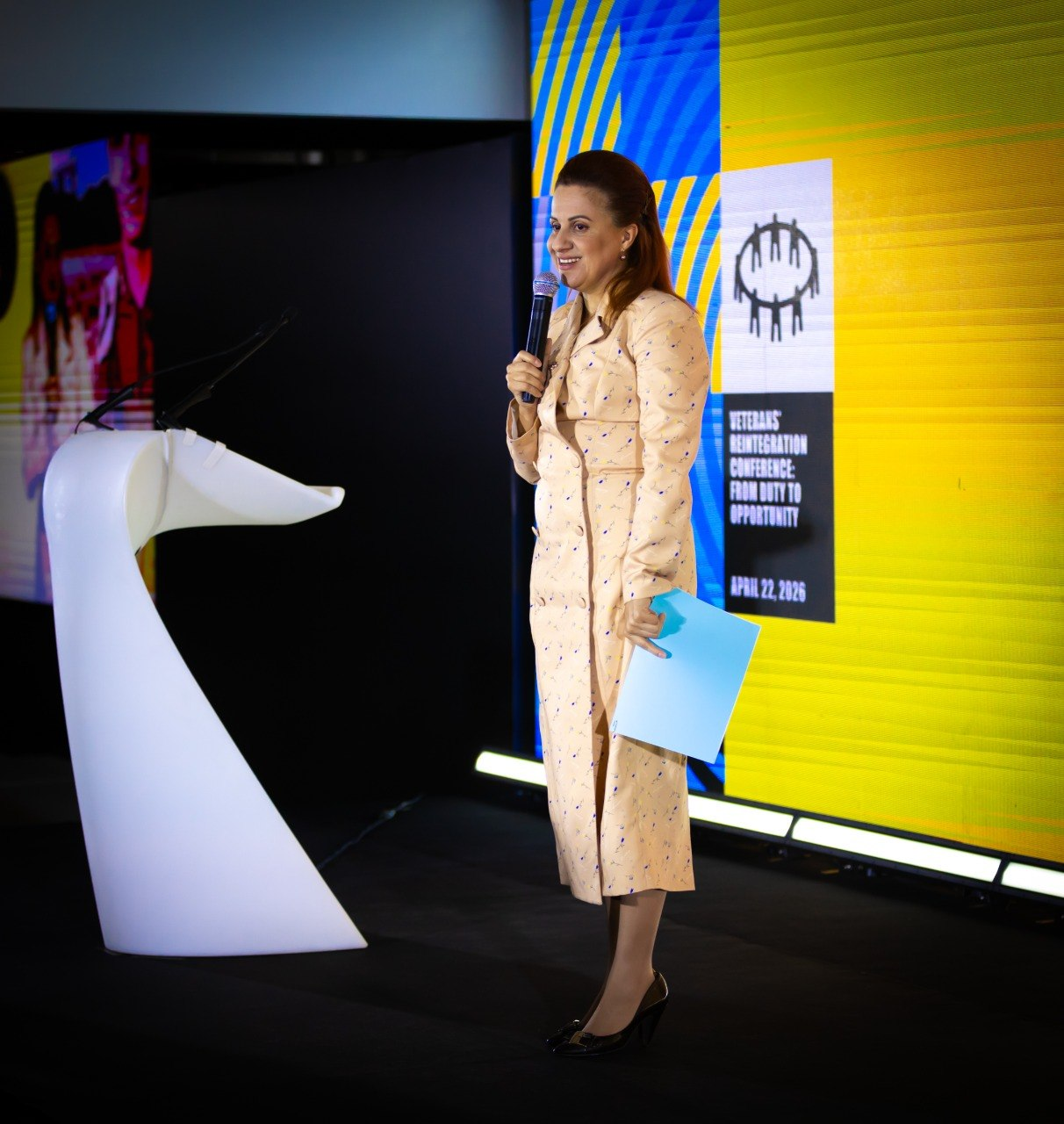 Жінка у світлому вбранні виступає з мікрофоном на сцені. Поруч із нею — біла трибуна футуристичної форми, а на задньому плані — яскравий екран із логотипом конференції «Veterans' Reintegration Conference» та датою 22 квітня 2026 року.