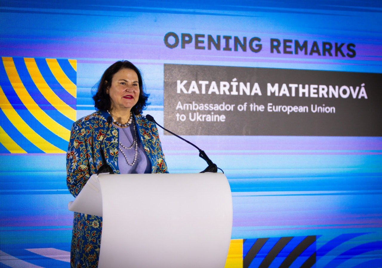 Жінка у жакеті з візерунком виступає за білою трибуною. На задньому плані — екран із написом «OPENING REMARKS» та інформацією про виступ Посла Європейського Союзу в Україні.