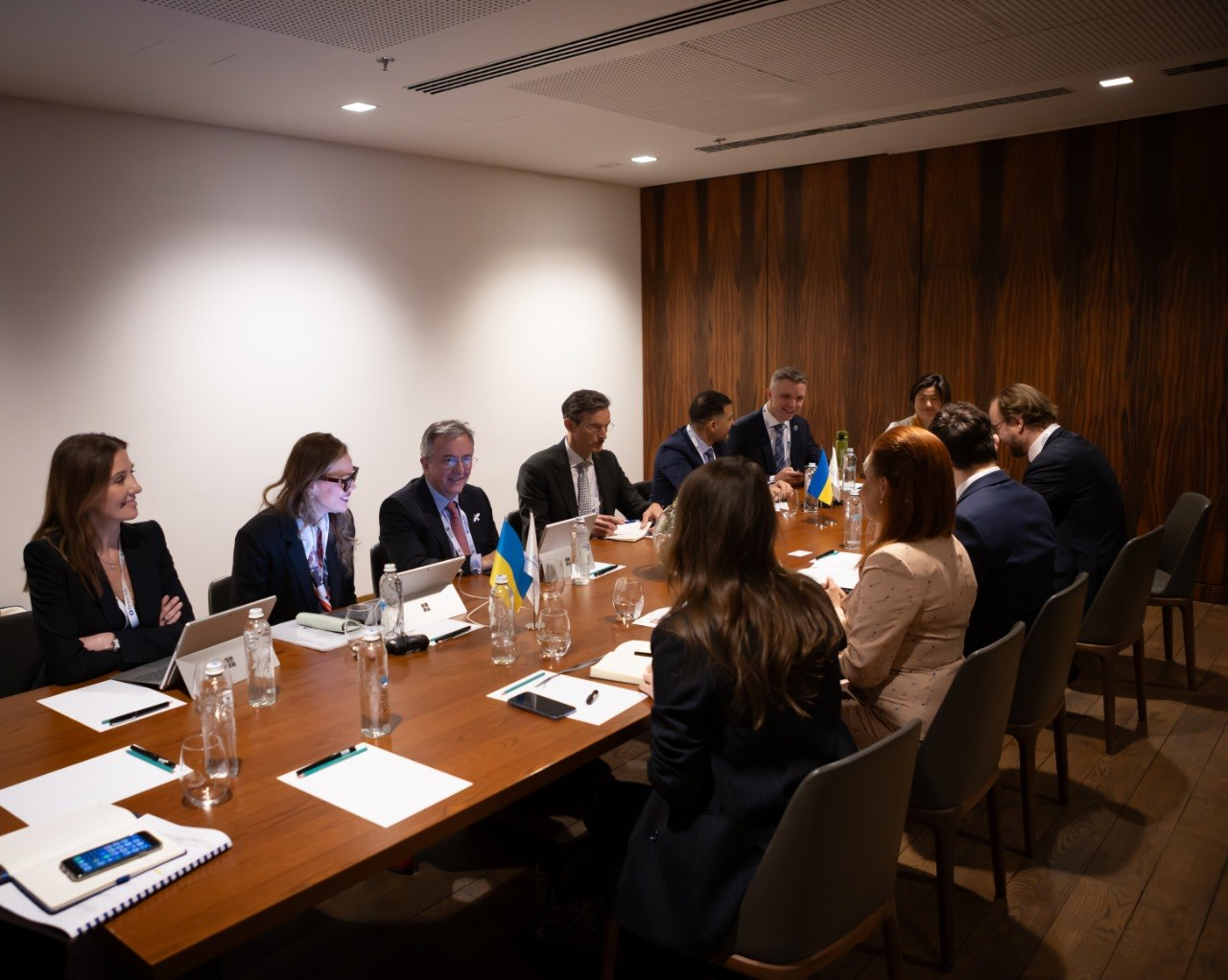 Група людей у ділових костюмах проводить зустріч за довгим дерев'яним столом у конференц-залі. На столі видно блокноти, ноутбуки та маленькі прапори України. Учасники спілкуються в офіційній, але доброзичливій атмосфері.