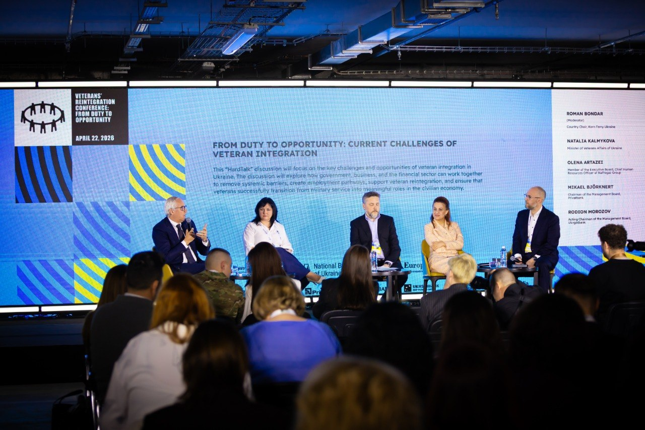 Панельна дискусія на конференції з реінтеграції ветеранів. П'ятеро спікерів сидять у кріслах на сцені перед великим екраном із написом «From Duty to Opportunity: Current Challenges of Veteran Integration». Перед сценою — аудиторія слухачів.