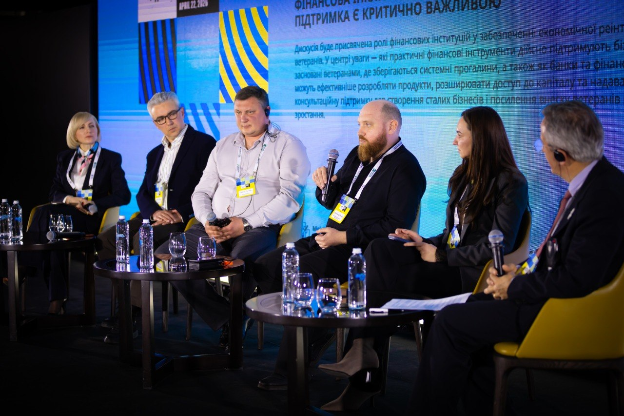 Група спікерів бере участь у панельній дискусії на тему фінансової підтримки ветеранів. Учасники сидять у кріслах на сцені, один із них виступає з мікрофоном. На задньому плані — великий екран із текстом про роль фінансових інституцій у реінтеграції.