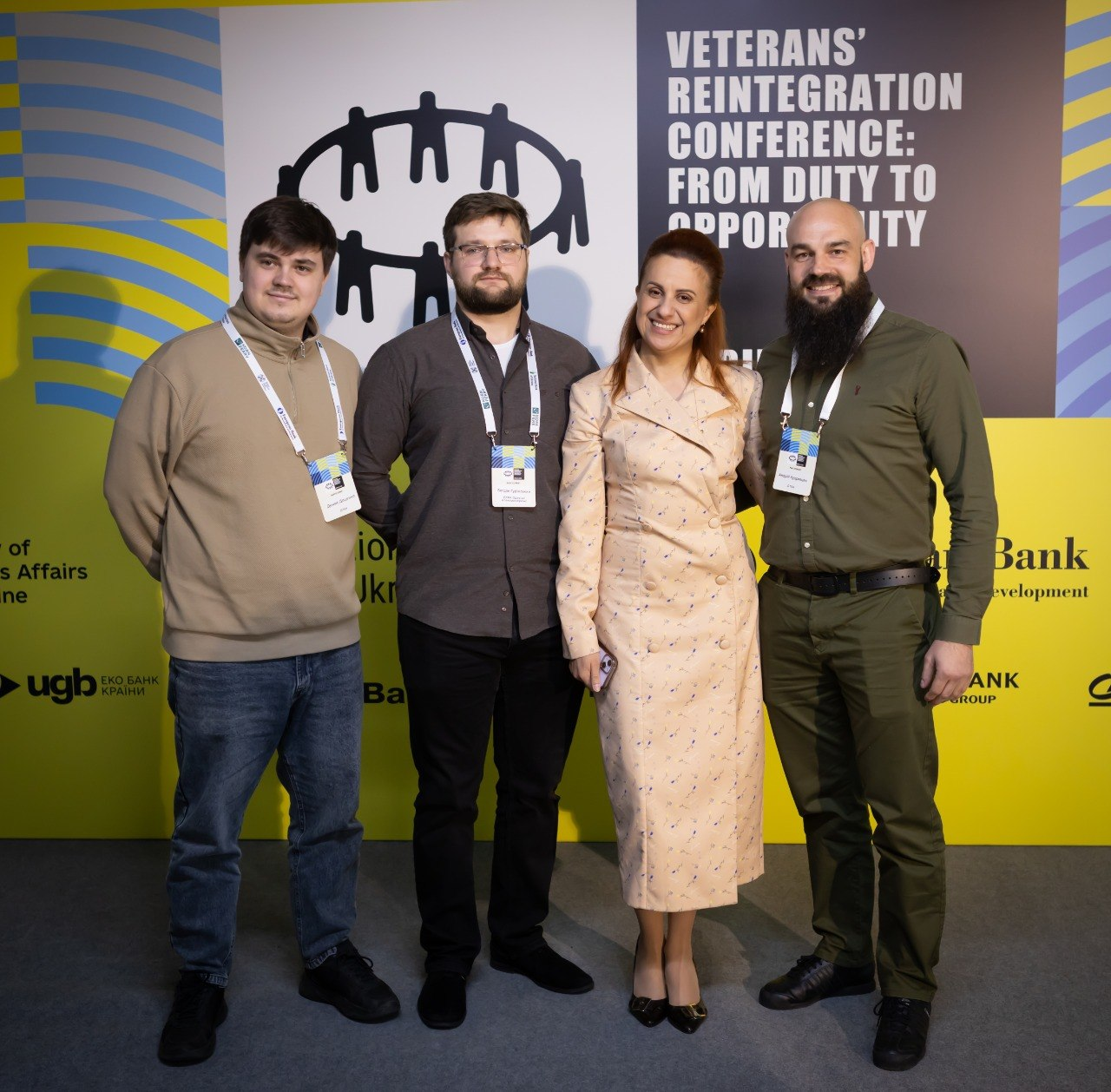 Група людей (три чоловіки та жінка) позують для фото на фоні банера конференції з реінтеграції ветеранів «Veterans’ Reintegration Conference». Усі учасники мають на шиї офіційні бейджі заходу.
