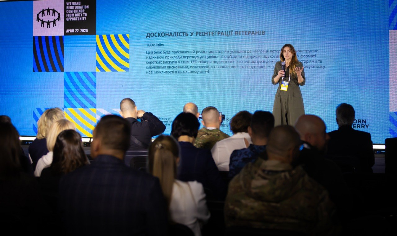 Жінка виступає на сцені перед аудиторією під час заходу у форматі TEDx. На великому екрані позаду неї заголовок «ДОСКОНАЛІСТЬ У РЕІНТЕГРАЦІЇ ВЕТЕРАНІВ» та опис блоку, присвяченого реальним історіям успіху ветеранів у цивільному житті.