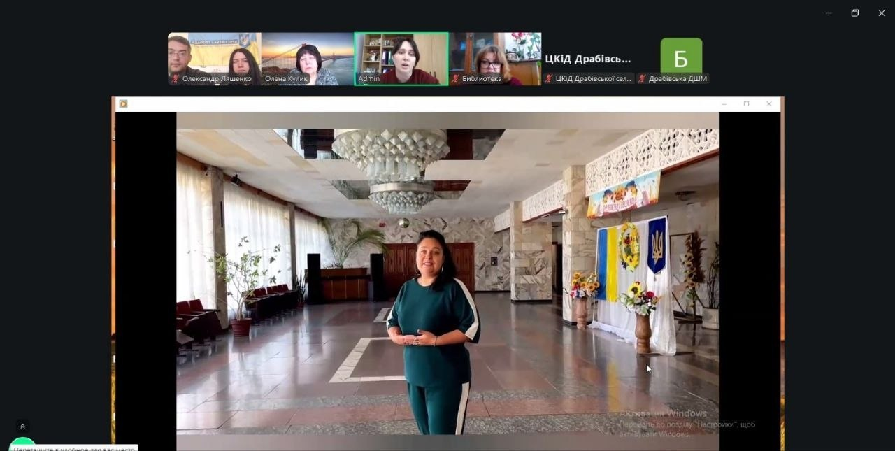 Скриншот онлайн-конференції у Zoom або Google Meet. У центральній частині екрана демонструється відео з жінкою, яка стоїть у просторому фоє з державною символікою України. У верхній частині — вікна з іншими учасниками зустрічі.