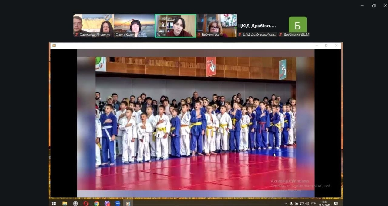 Скриншот онлайн-зустрічі, на якому демонструється фотографія великої групи дітей у спортивній формі для єдиноборств (кімоно). Діти стоять у шерензі на татамі, поклавши руку на серце. У верхній частині екрана видно вікна з іншими учасниками відеоконференції.