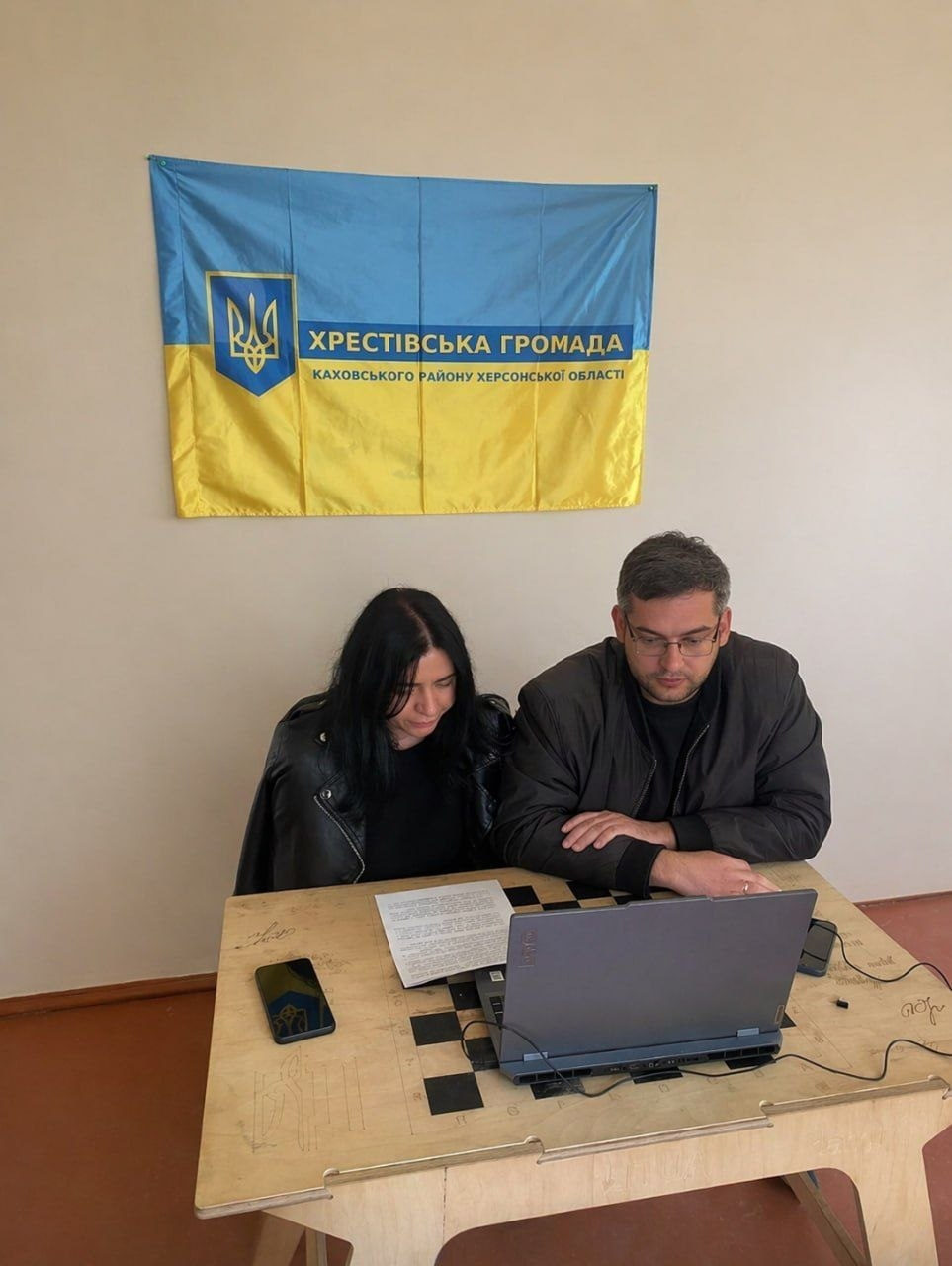 Чоловік та жінка працюють за ноутбуком у приміщенні. На стіні позаду них висить прапор Хрестівської громади Каховського району Херсонської області. На столі поруч із ноутбуком лежать документи та мобільний телефон.