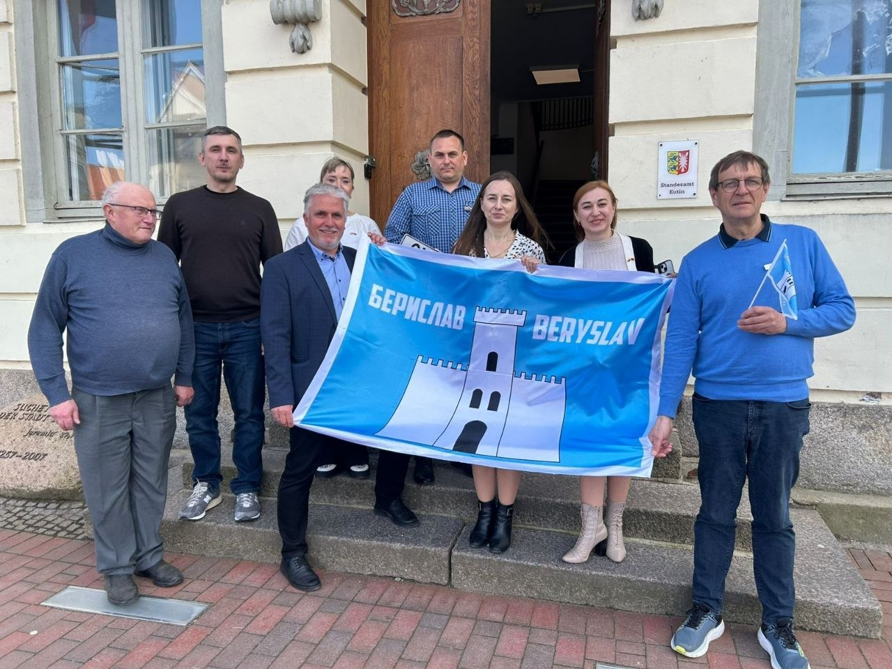 Група людей стоїть на сходах будівлі, тримаючи великий блакитно-білий прапор із написом «БЕРИСЛАВ / BERYSLAV» та зображенням фортечної вежі. Учасники зустрічі позують для спільного фото в офіційній обстановці.