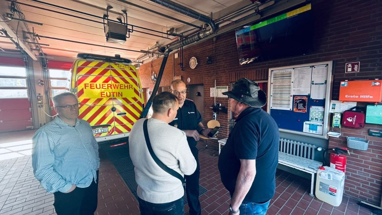Група чоловіків спілкується в приміщенні пожежної частини поруч із рятувальним автомобілем «FEUERWEHR EUTIN». На стіні видно інформаційний стенд, аптечку першої допомоги та вогнегасник.