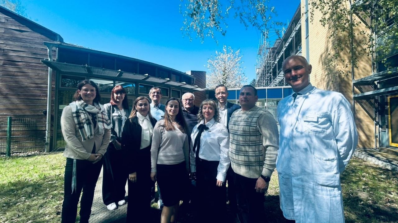 Група людей позує для спільного фото на подвір’ї сучасної будівлі у сонячний день. Один із чоловіків на передньому плані одягнений у білий медичний халат. На фоні видно зелені насадження та фасад споруди в процесі реновації.