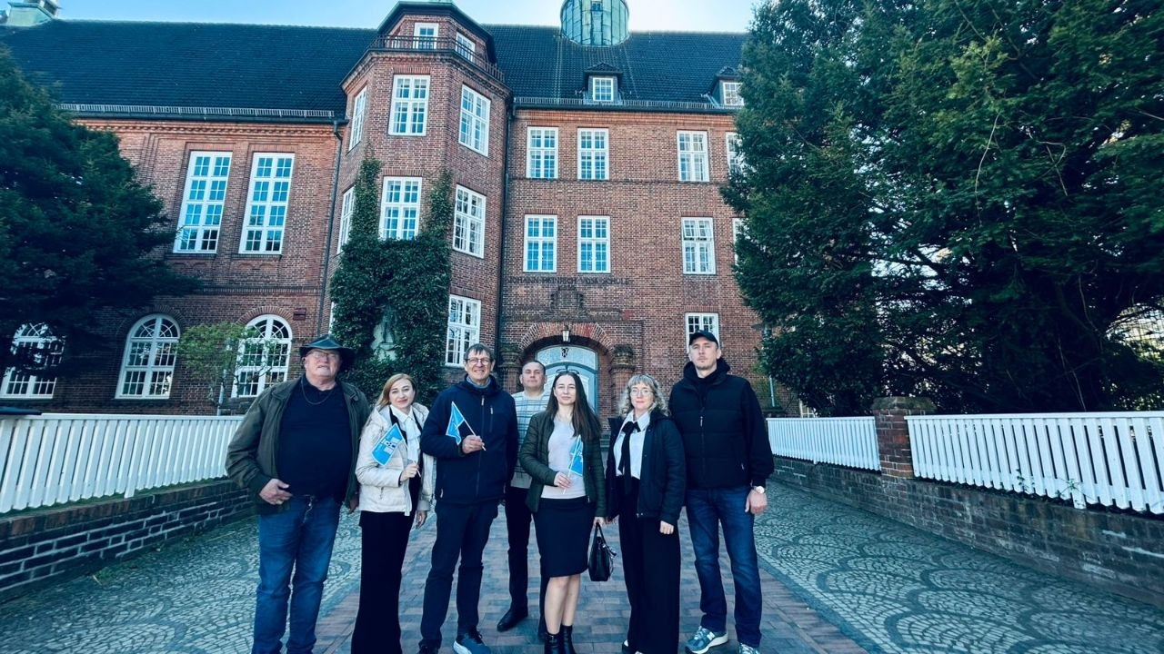 Група людей позує для фото перед великою триповерховою будівлею з червоної цегли в класичному європейському стилі. Дехто з учасників тримає в руках невеликі блакитні прапорці. Будівлю оточують високі дерева та низький білий паркан.